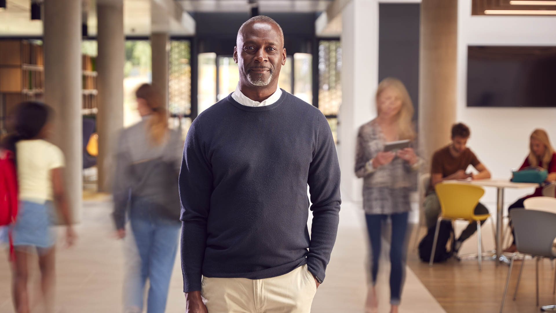 older african american male teacher standing in front of a college common area with students walking by