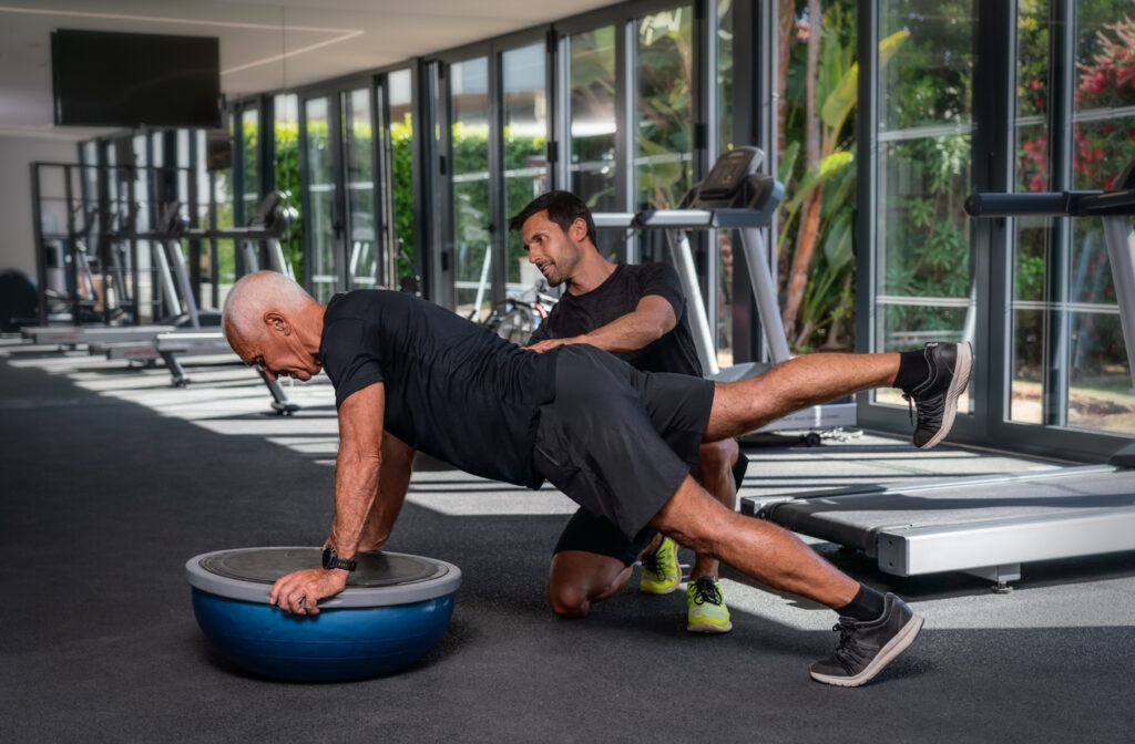 An elderly Caucasian man, in the gym, with a personal fitness trainer, in rehabilitation, doing exercises for balance on a gymnastic ball. High quality photo